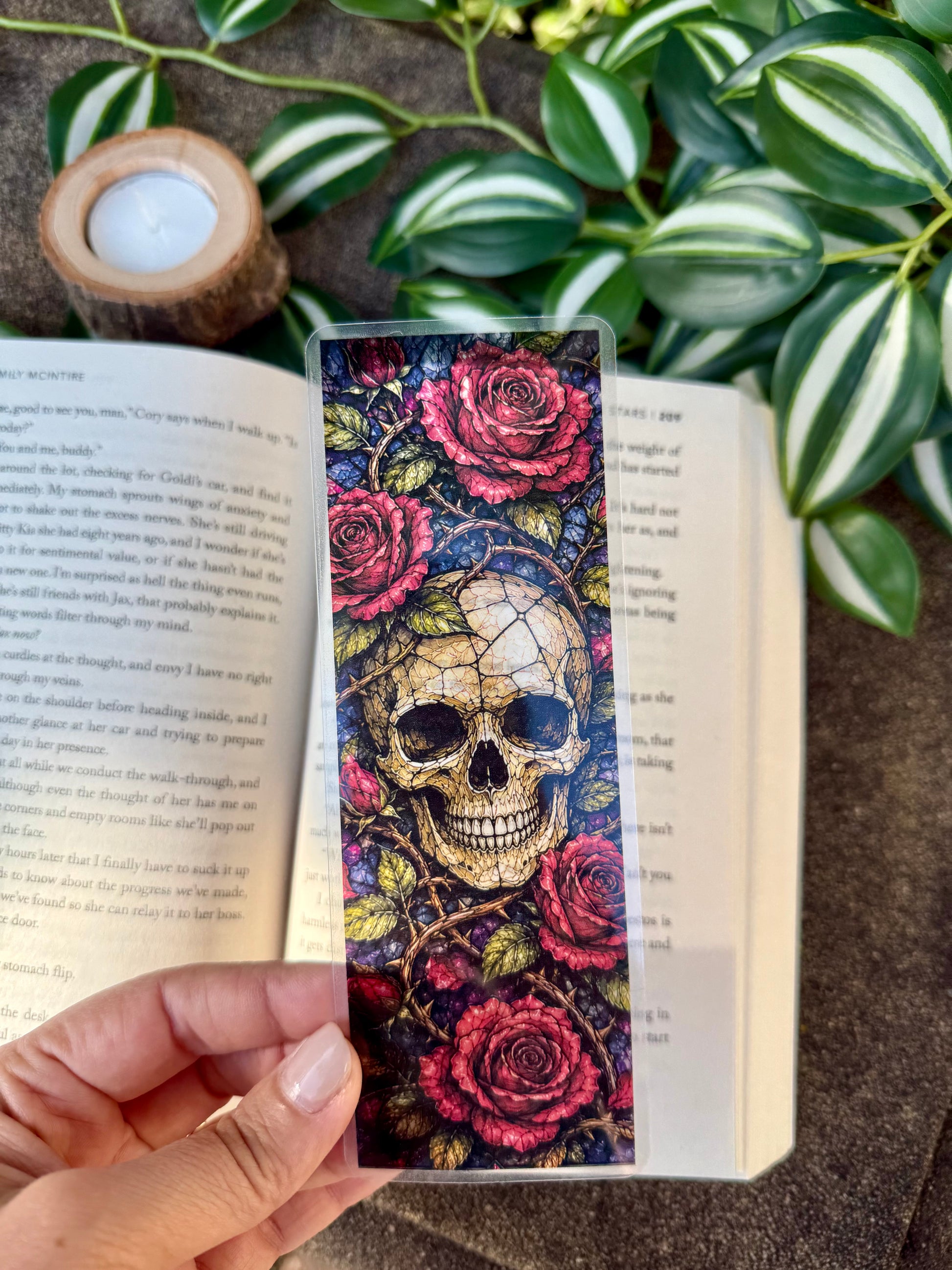 Colorful stained glass skull and rose bookmark held over an open book with plants in the background