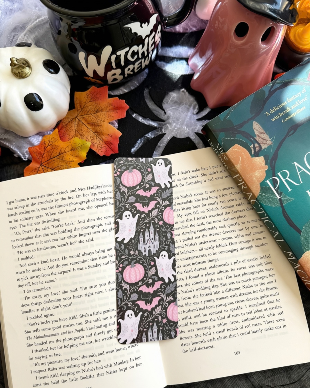 A floral bookmark of ghosts, castle, pumpkins on a black background. The bookmark is placed on an open book surrounded by Halloween decorations. 