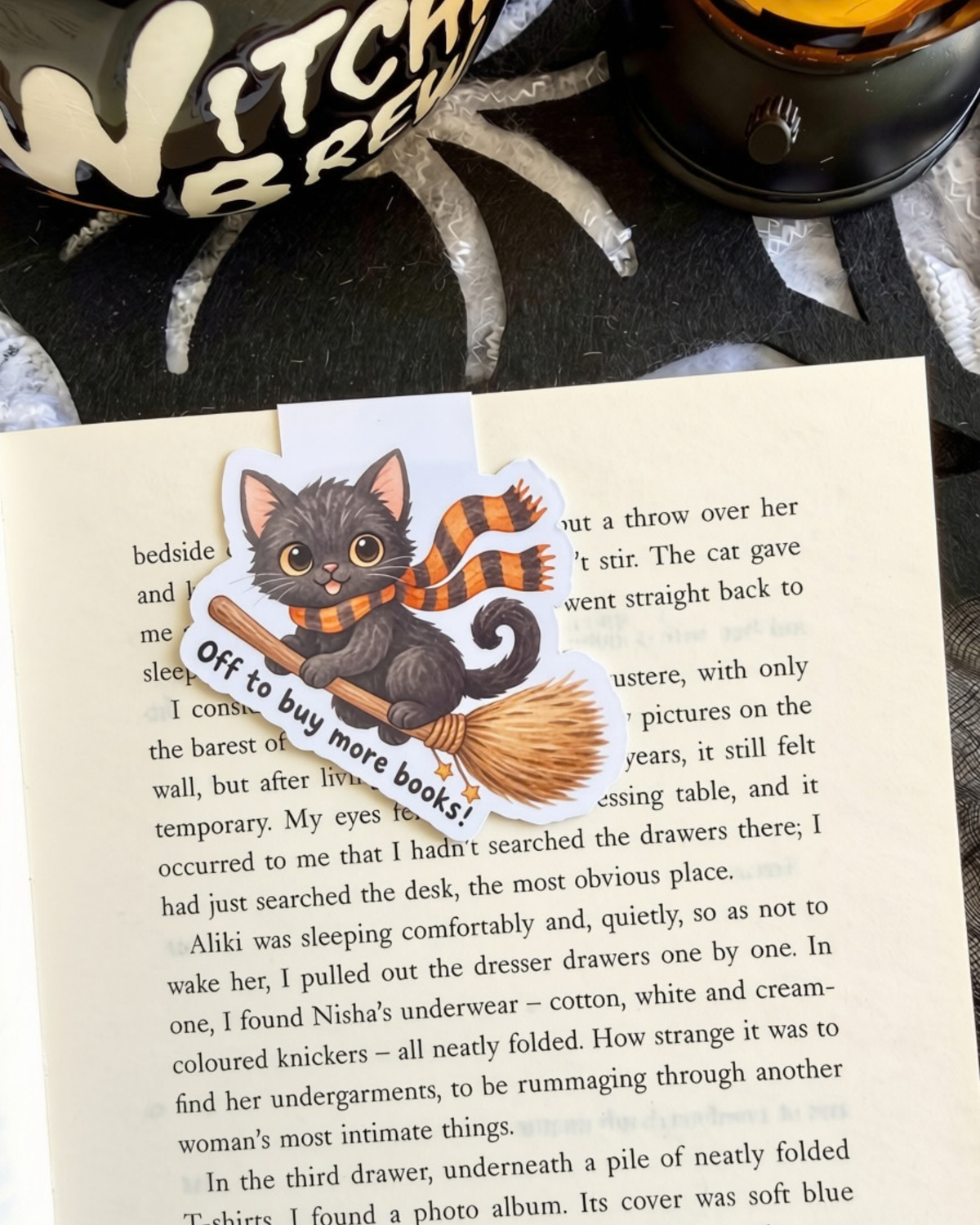 A magnetic bookmark of a black cat wearing an orange and black striped scarf and a brown broomstick, placed on an open book page. The sticker has the text 'Off to buy more books!' on a white banner.