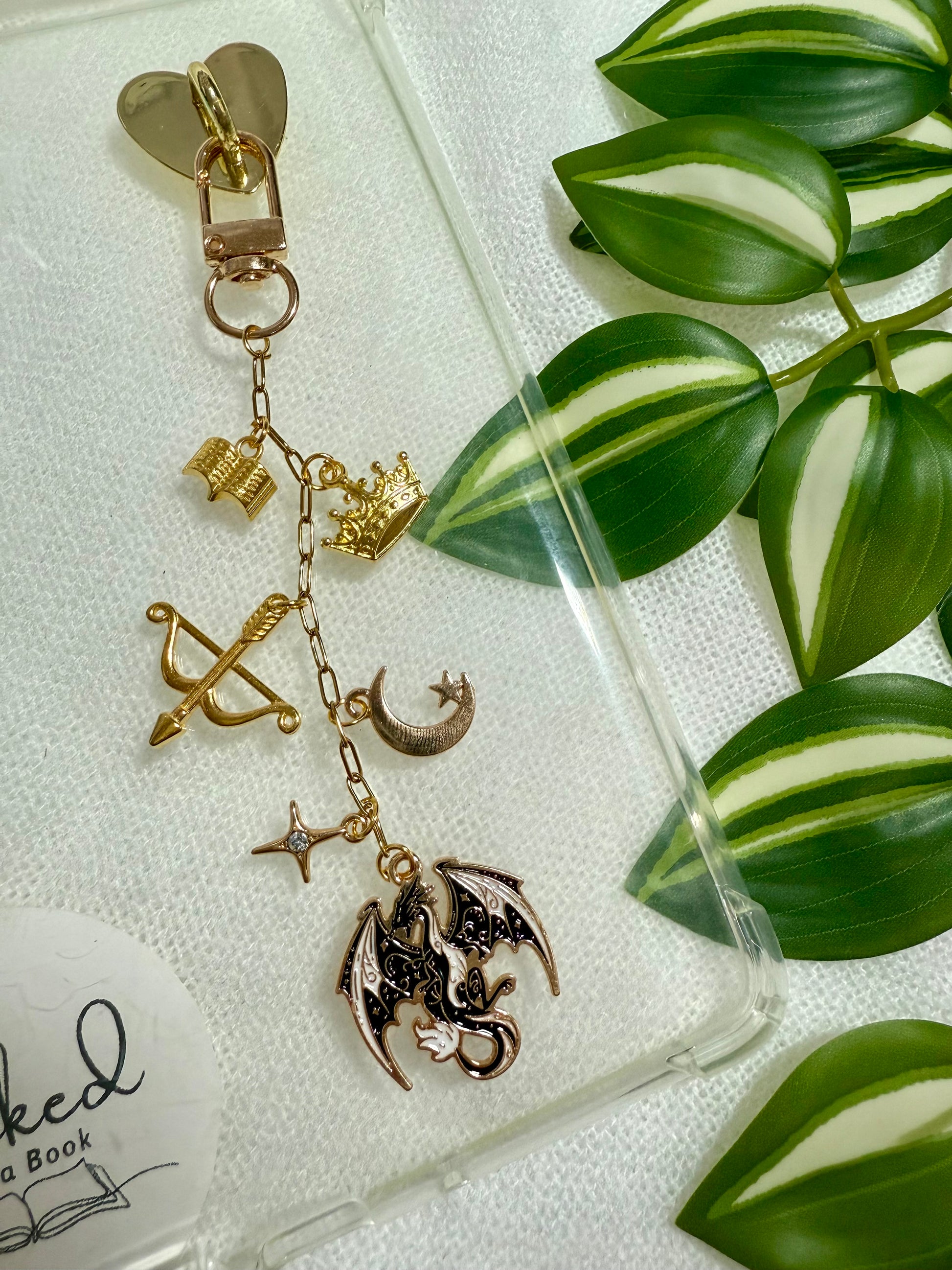 A chain with gold fantasy charms such as a dragon, crown, bow and arrow attached to a clear kindle case.