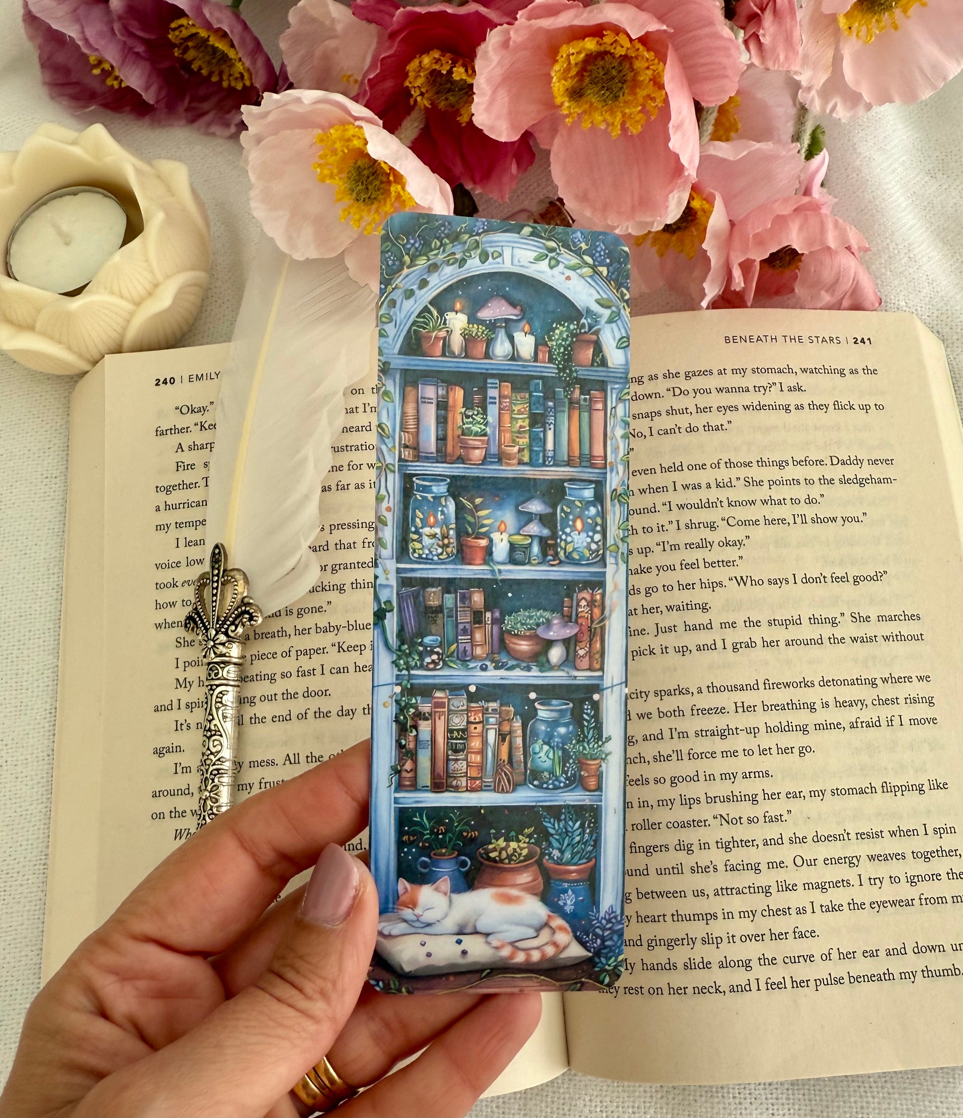 Hand holding a bookmark with a bookshelf design over an open book with flowers and a candle in the background.