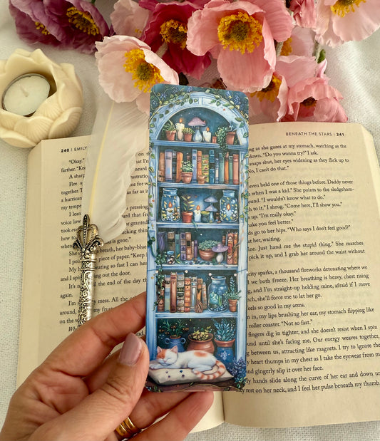 Hand holding a bookmark with a bookshelf design over an open book with flowers and a candle in the background.