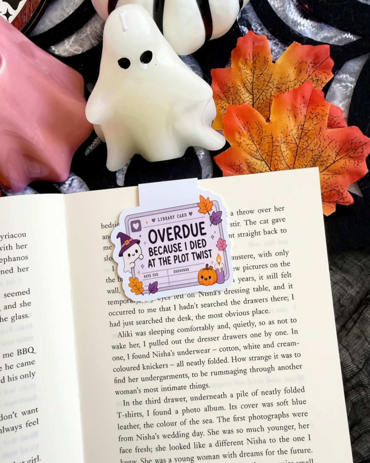An open book spread showing a magnetic bookmark cartoon witch illustration and the text 'OVERDUE BECAUSE I DIED AT THE PLOT TWIST'. The scene is styled with pink roses, white flowers, and orange autumn leaves on a dark background.