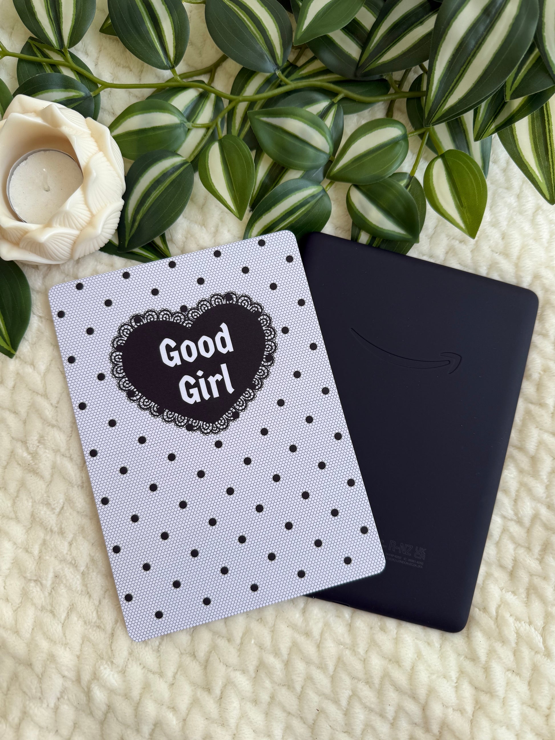 A black Kindle e-reader lying on a cream knit blanket, styled with a black-and-white polka dot notebook that says 'Good Girl' in a heart, green striped leaves, and a white rose.