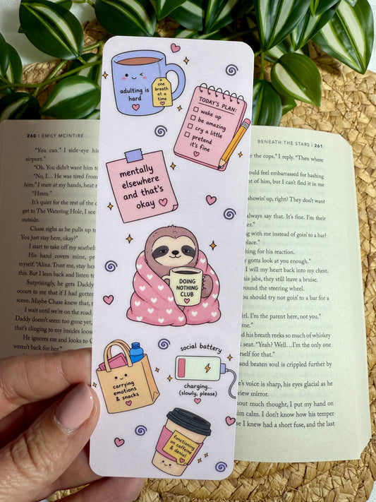 A hand holds a long white bookmark or sticker sheet featuring cute cartoon illustrations, including a sloth, coffee cup, notebook, and shopping bag, placed over open book pages with green leaves in the background.