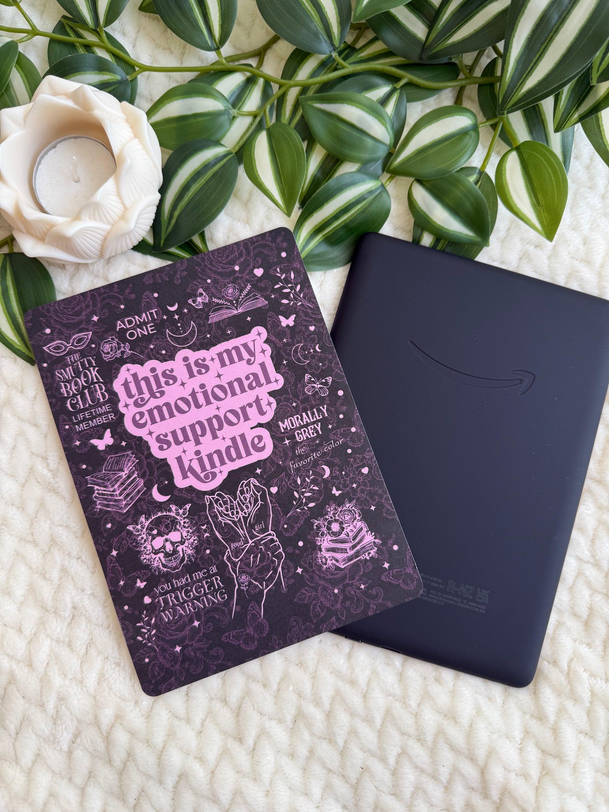A dark purple illustrated notebook with pink text reading 'this is my emotional support kindle' is placed on top of a black Kindle e-reader, styled on a cream textured blanket with green leaves and a white flower.