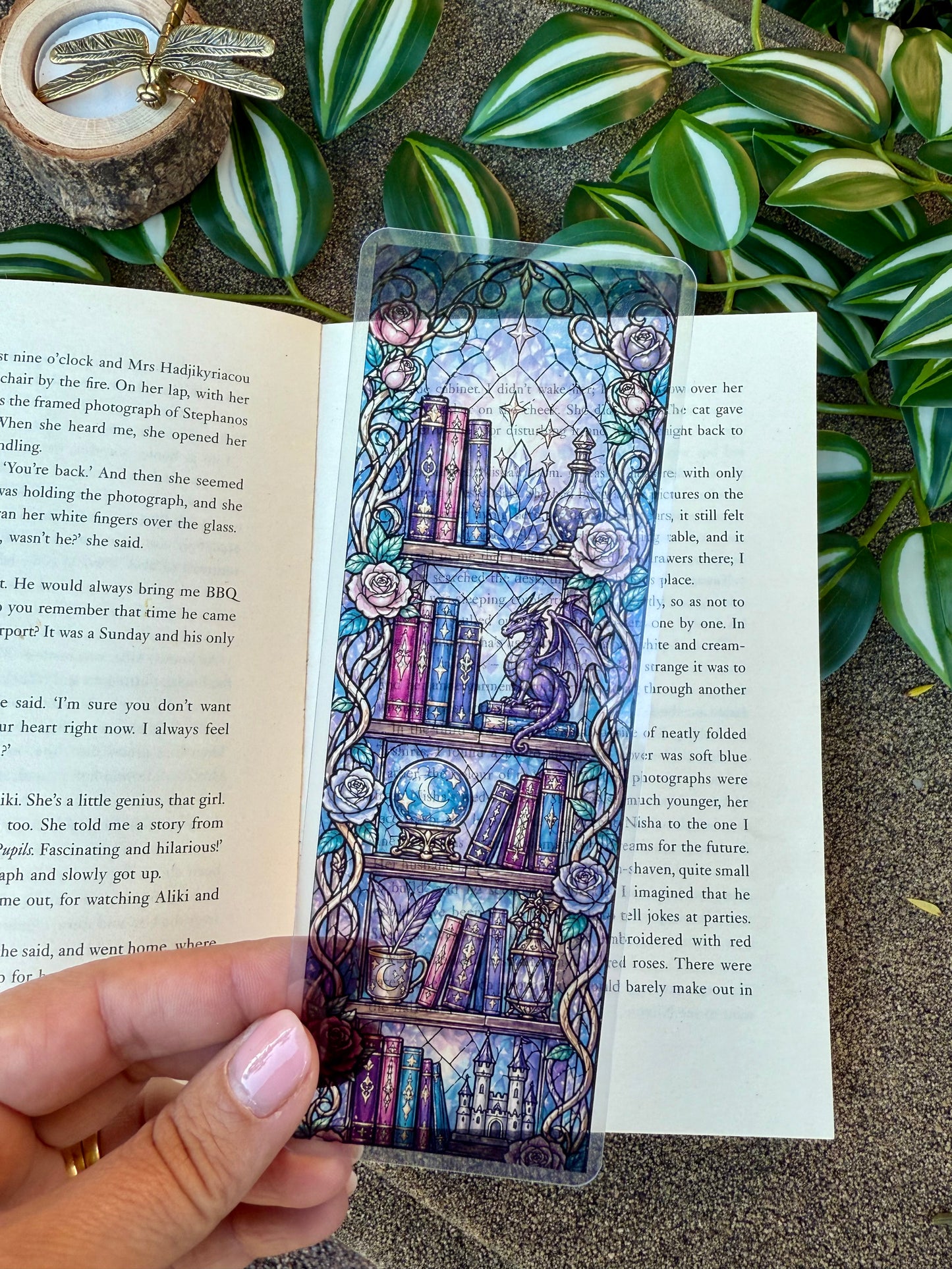 A transparent stained glass bookmark of a bookshelf with fantasy elements such as a dragon and crystals. 