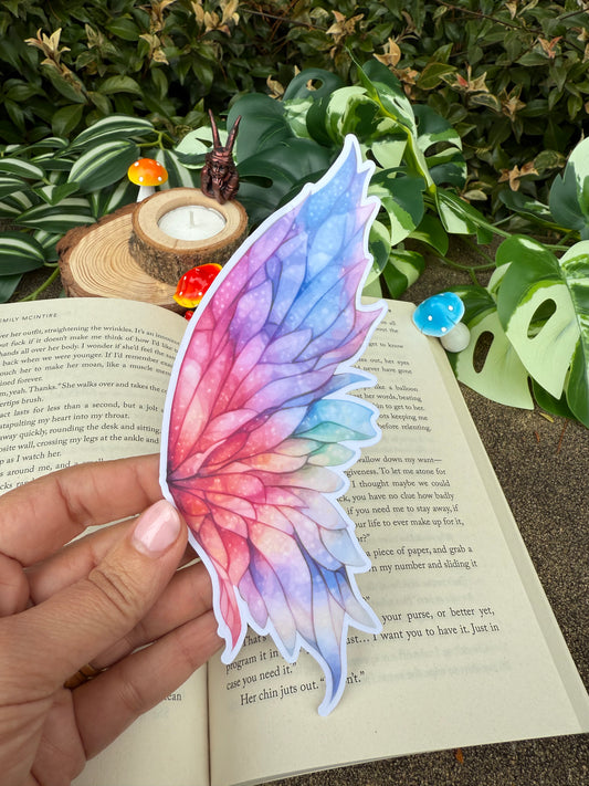 A hand holds a die-cut fairy wing bookmark with a watercolor design in shades of pink, purple, and blue, positioned over an open book in a garden-themed setting with small mushrooms and green leaves.