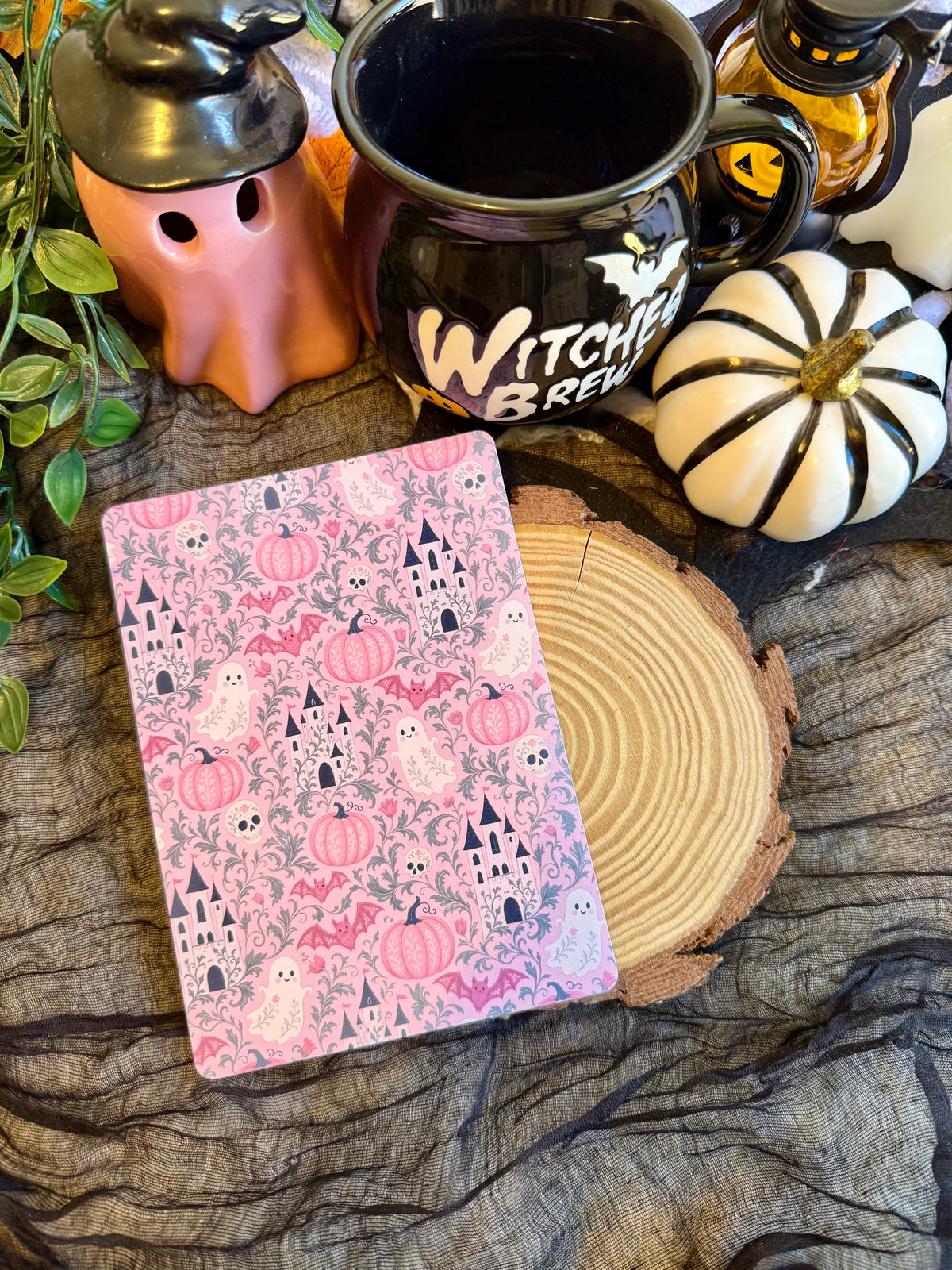 A kindle insert witch-themed design of pumpkins, castles, and ghosts, styled on a rustic wood slice next to a black 'Witch Brew' mug, a ghost-shaped ceramic, and a small white pumpkin.