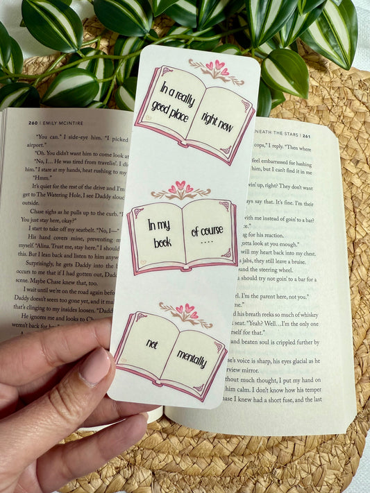 Three decorative bookmarks shaped like open books with pink floral accents and printed phrases, shown being held over an open book on a woven placemat with green leaves in the background.
