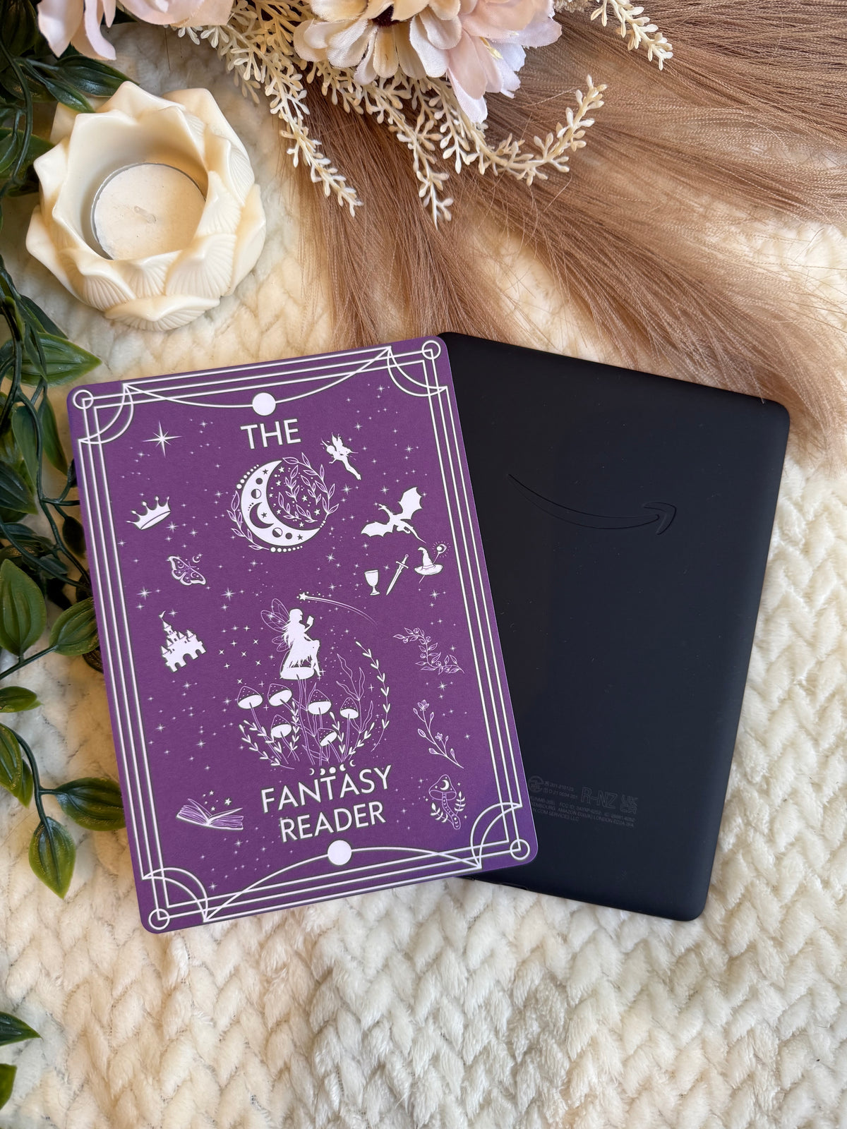 A purple kindle insert labeled 'THE FANTASY READER' with white fantasy illustrations, placed beside a black kindle on a cream textured blanket with dried flowers and greenery around it.