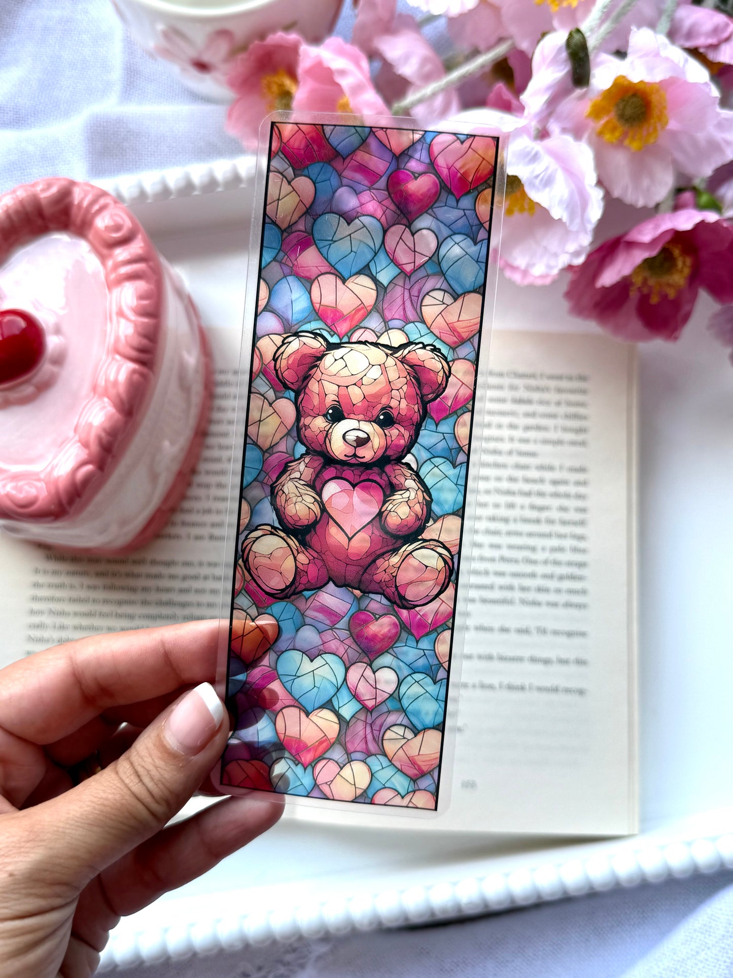 Colorful teddy bear and hearts stained glass bookmark held over an open book with flowers and a pink candle in the background.