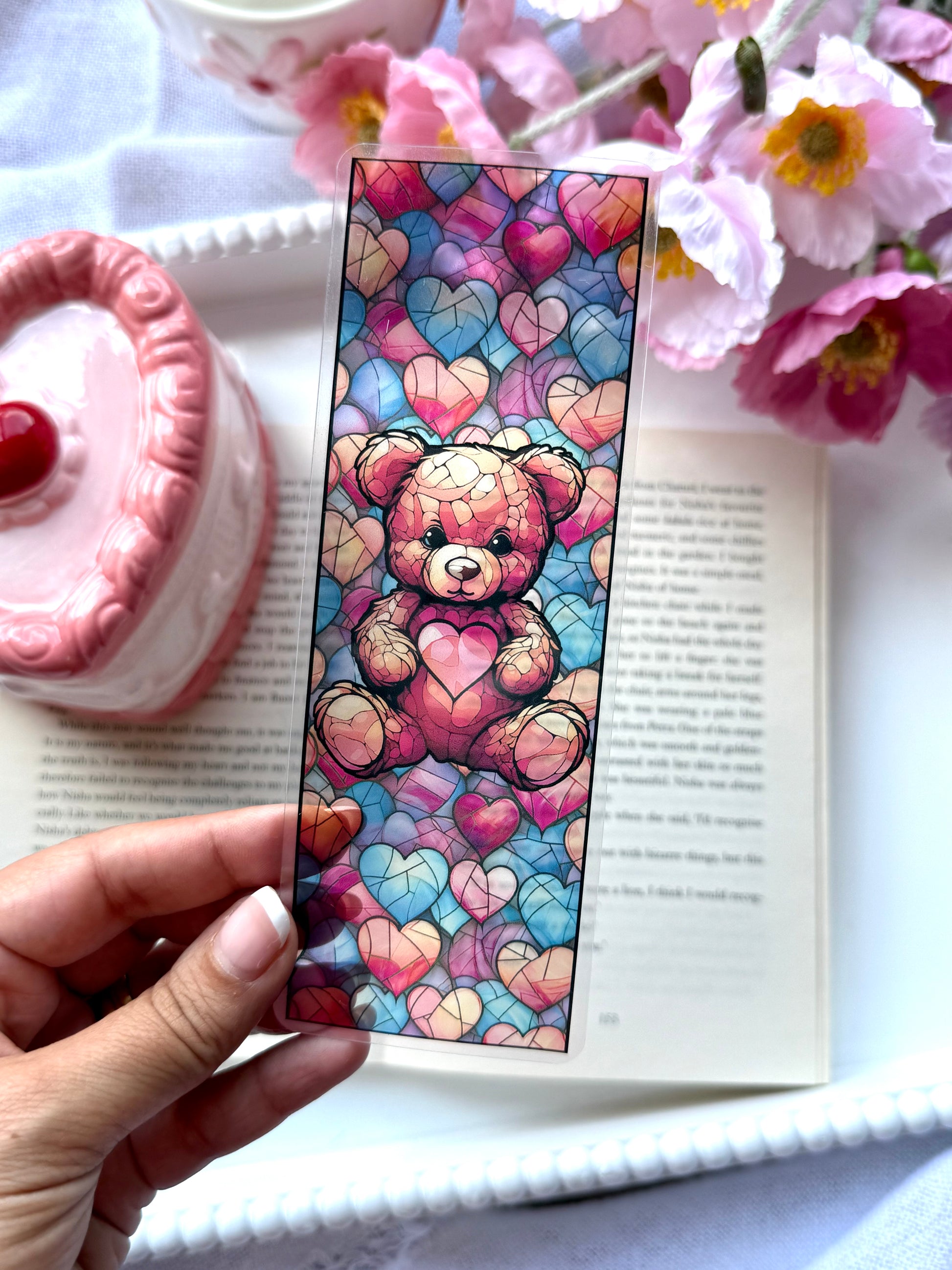 Colorful teddy bear and hearts stained glass bookmark held over an open book with flowers and a pink candle in the background.
