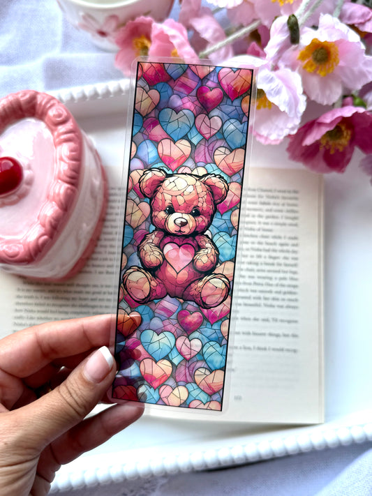 Colorful teddy bear and hearts stained glass bookmark held over an open book with flowers and a pink candle in the background.