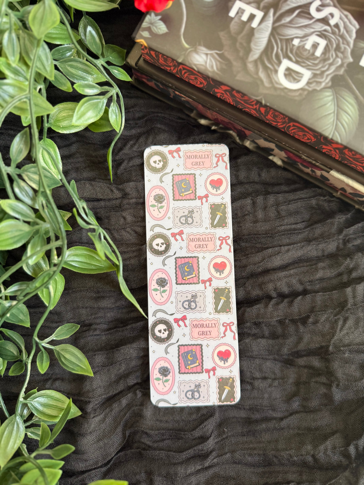 A slim rectangular card or bookmark with a vintage gothic illustration pattern featuring skulls, roses, and hearts, displayed on a dark textured fabric next to green leaves and a decorative book.