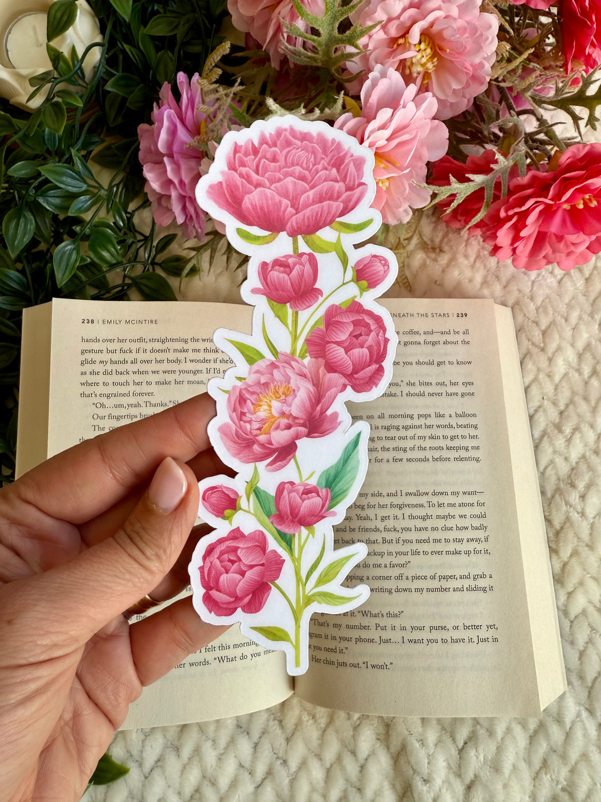 A hand holds a die-cut bookmark featuring pink peony flowers with green leaves over an open book, with a background of dried pink flowers.