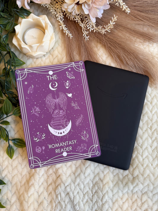 A kindle insert 'THE ROMANTASY READER' in purple with white line art, placed next to a black kindle on a cream knit blanket with decorative dried flowers, greenery, and a candle.