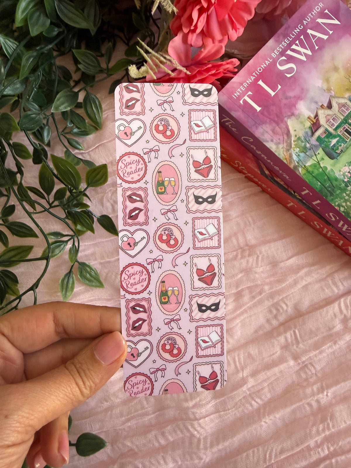 A hand holds a long rectangular bookmark or card featuring a pink and white pattern of lips, hearts, and decorative frames, styled with a pink textured fabric background, artificial green leaves, and a book in the corner.