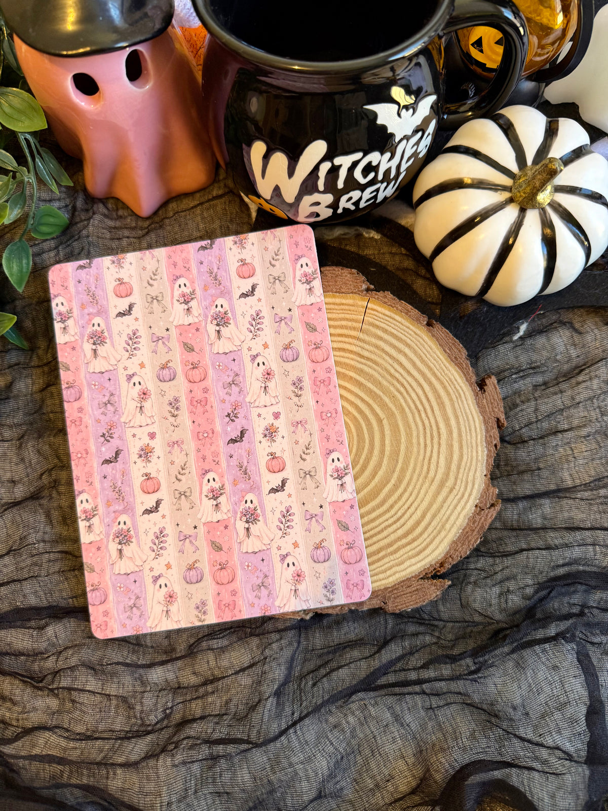 A kindle insert with a pink and beige Halloween-themed washi tape pattern featuring pumpkins, ghosts, and bats, styled on a rustic wood slice with a black Witch Brew mug, a white pumpkin, and a ghost figurine.