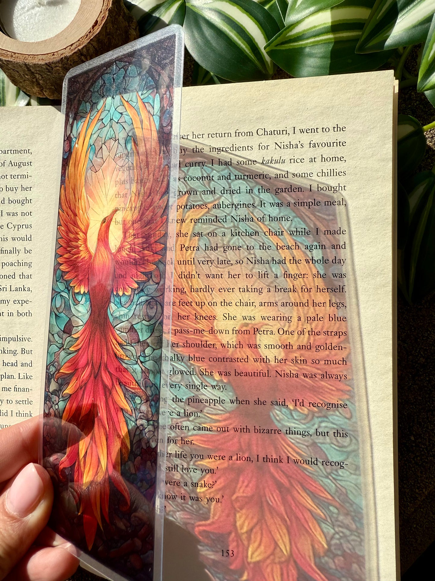 A transparent stained glass style bookmark of a phoenix. The bookmark is placed against a page of a book allowing the light to filter through the bookmark and project the image on the page.