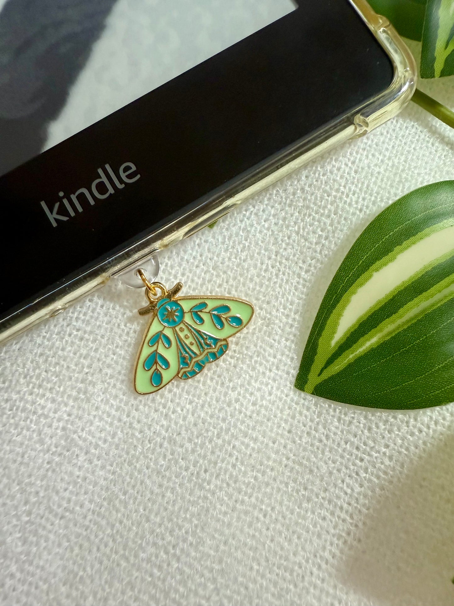 Green enamel moth pendant on a white surface with a Kindle device in the background