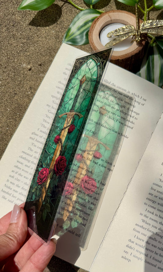 Decorative stained glass bookmark with a sword and roses design with an emerald green background held over an open book showing the light reflecting onto the book from the bookmark.