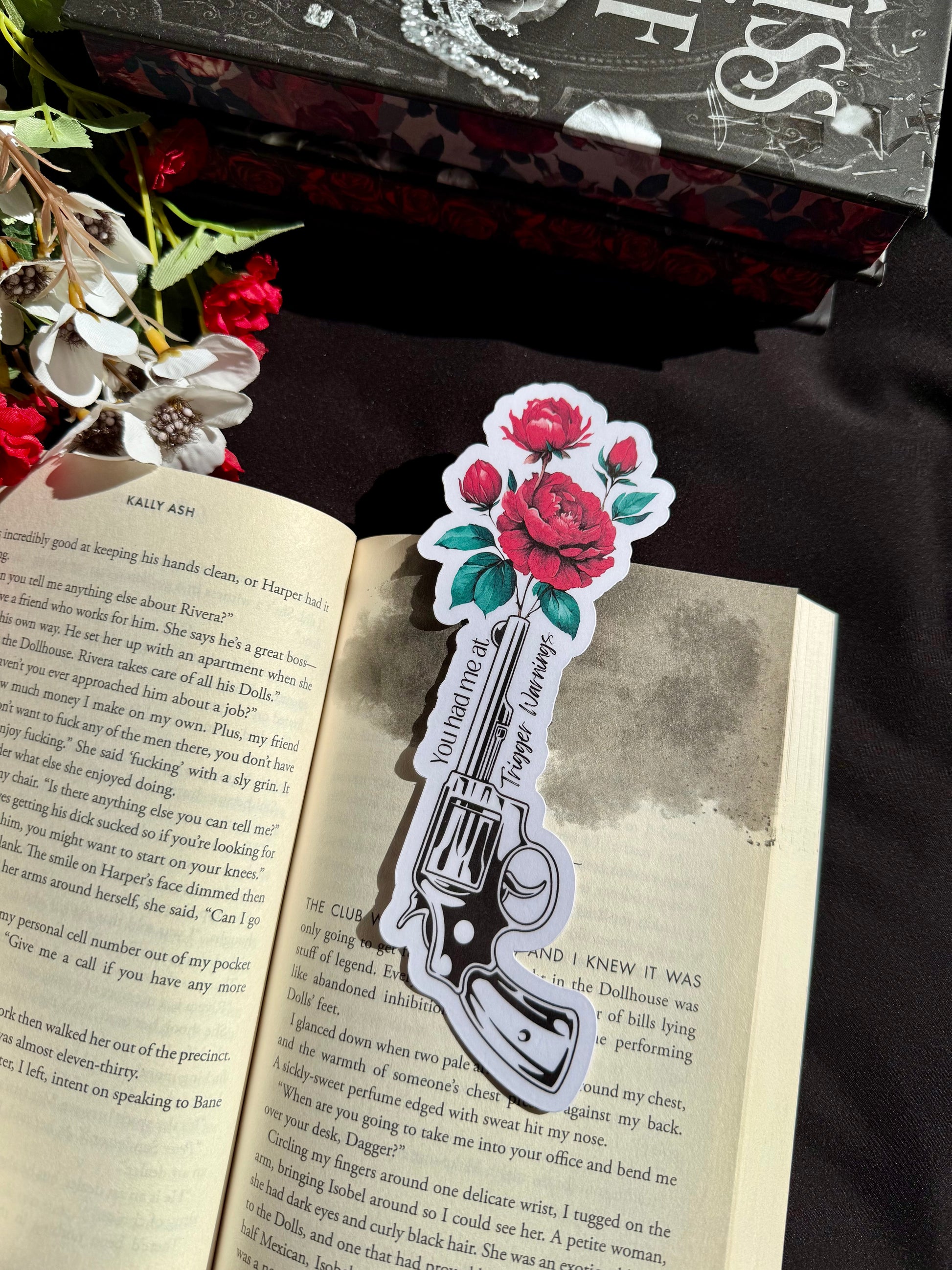 A die cut bookmark of a gun and roses sticking out of the gun, with the words You had me a trigger warnings. The bookmark is on an open book.