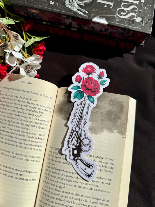 A die cut bookmark of a gun and roses sticking out of the gun, with the words You had me a trigger warnings. The bookmark is on an open book.