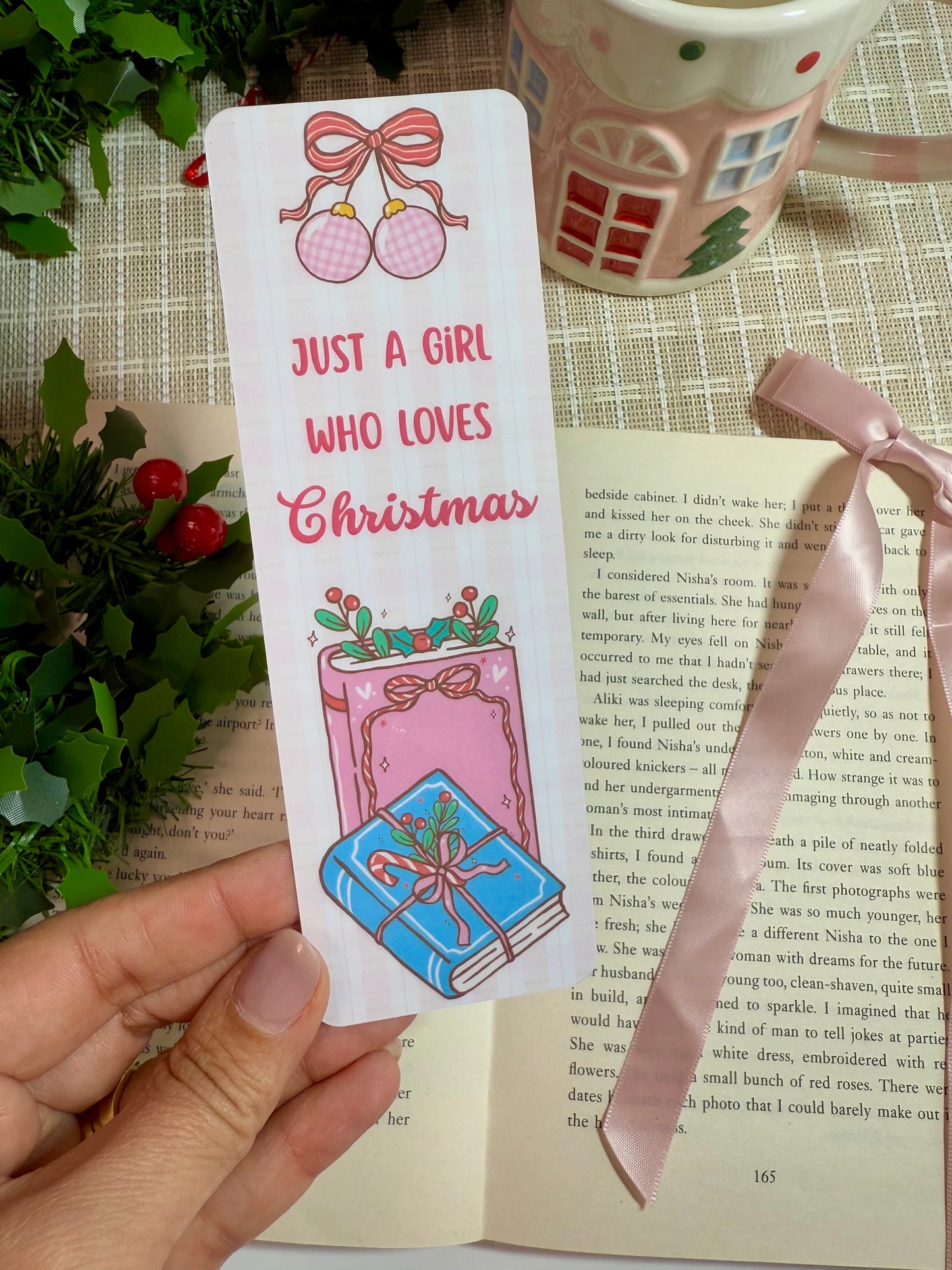 Bookmark with Christmas design held over an open book, with a mug and greenery in the background.