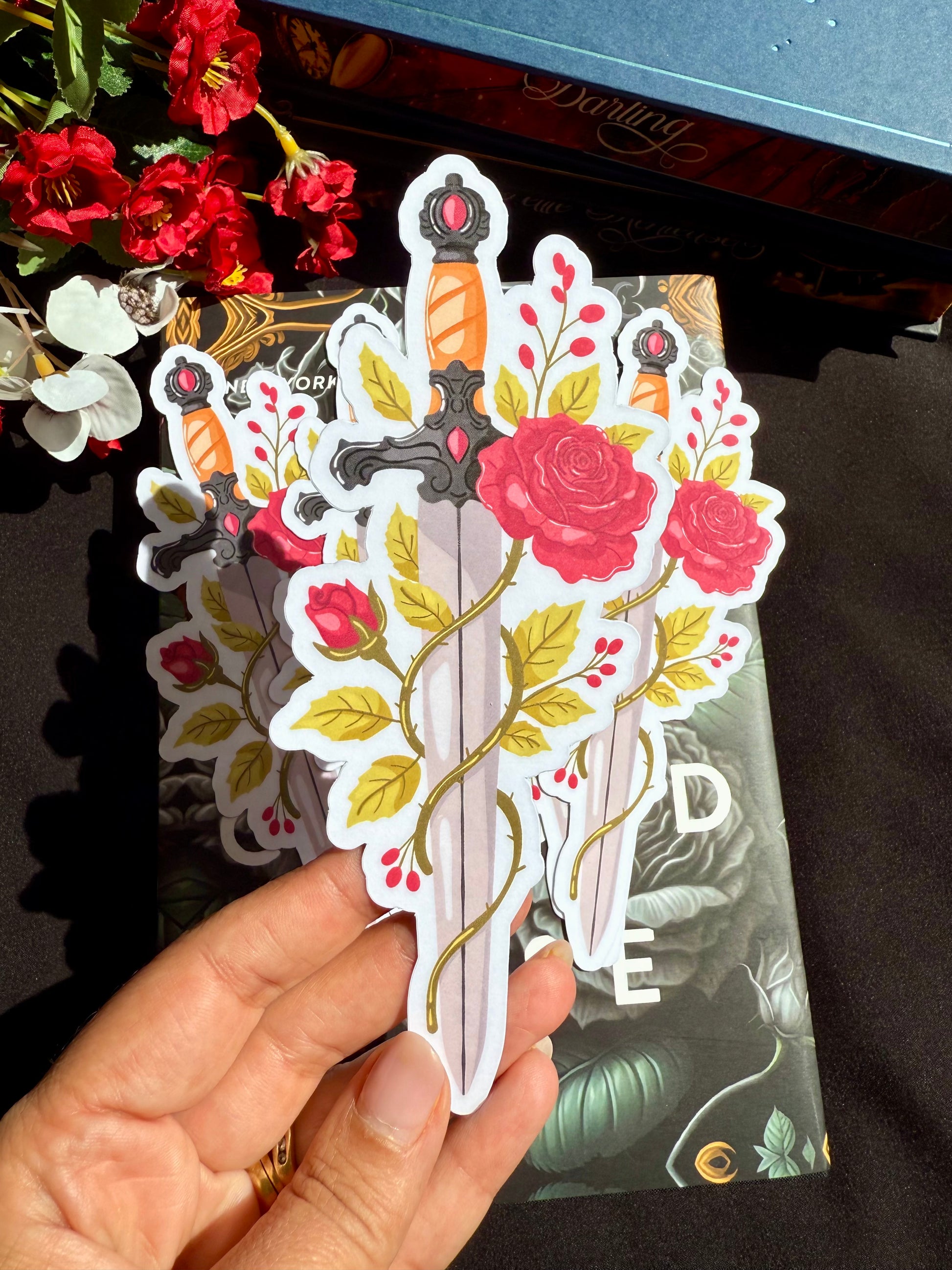 A die cut bookmark of a sword with red roses wrapped around it. The bookmark is on a dark romance book.