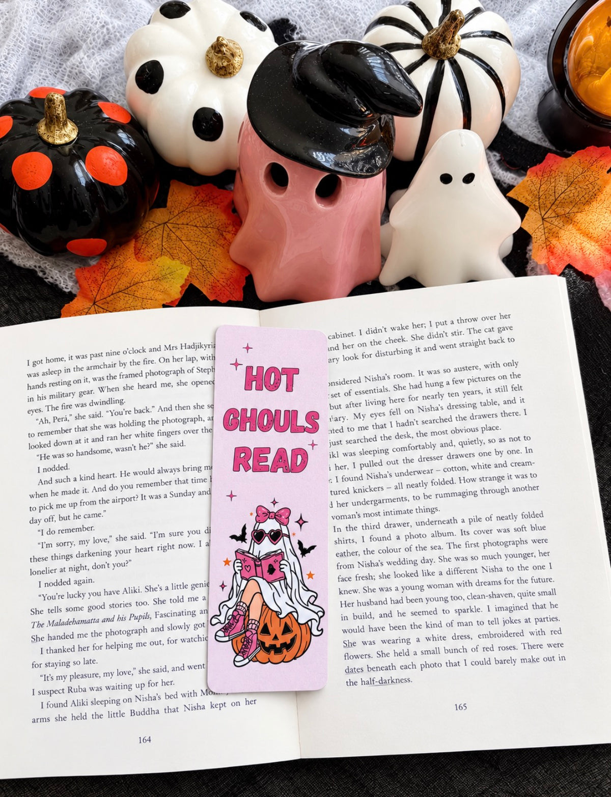 An open book spread laid on a white surface, featuring a pink bookmark with the title 'HOT GHOULS READ' and a cartoon ghost illustration. Surrounding the book are autumn leaves, a small pumpkin, a black hat, and a pink piggy bank.