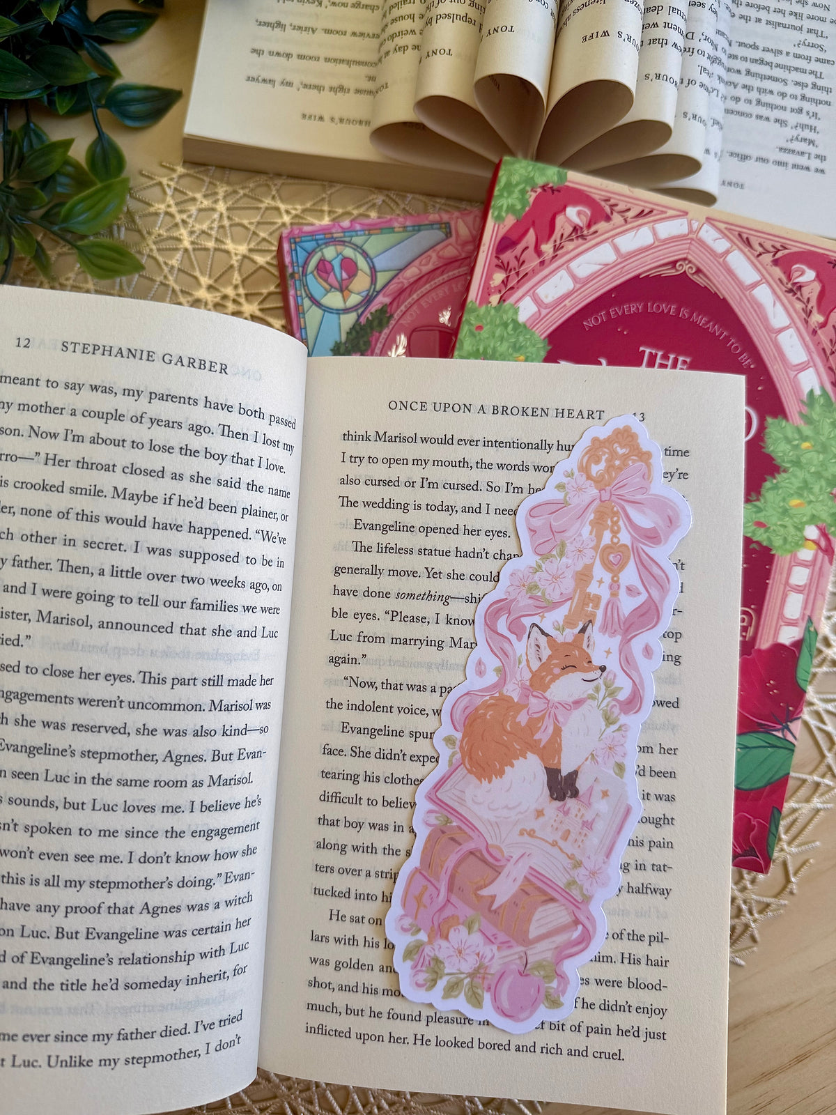 A die cut bookmark of a fox sitting on a stack of books. the bookmark is placed on an open book