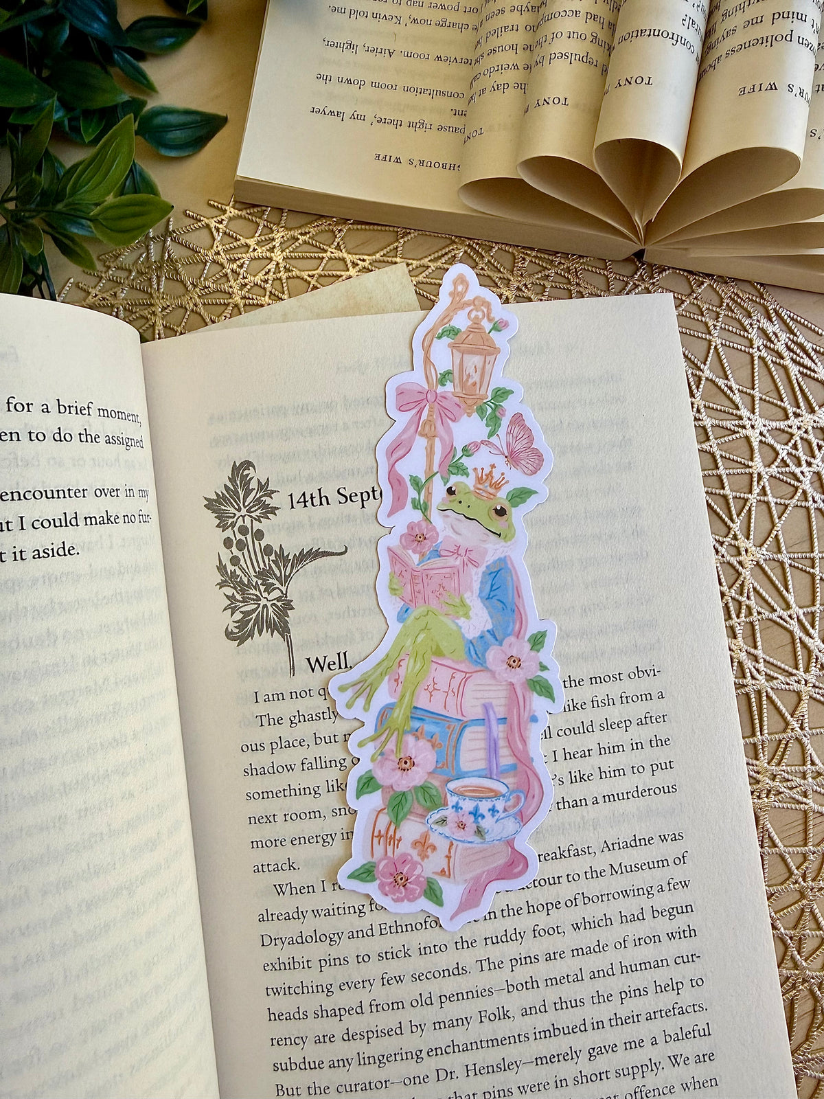A die cut bookmark of a frog sitting on a stack of books. the bookmark is placed on an open page of a book.