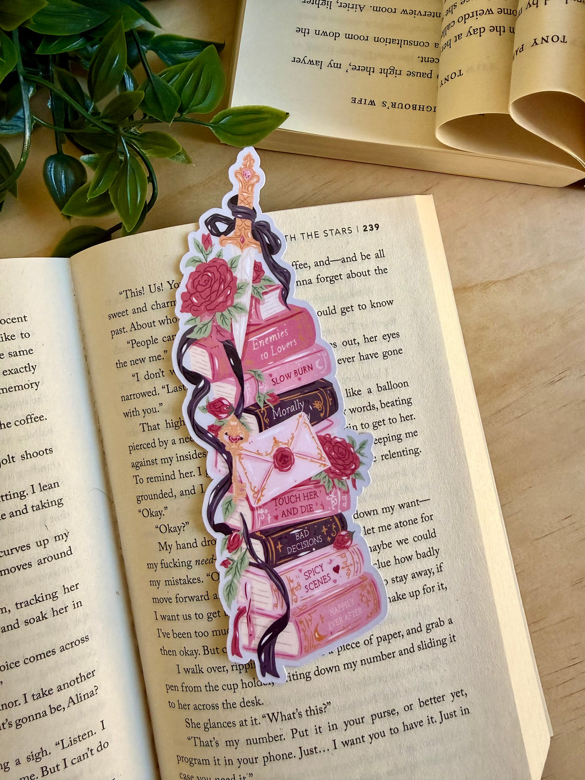 A die cut bookmark of a book stack with dark romance tropes and a sword on top. The bookmark is placed on an open book with foliage in the background.